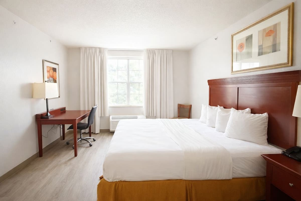 A hotel room with a large bed, a desk with a chair, a window with curtains, and a framed picture on the wall.