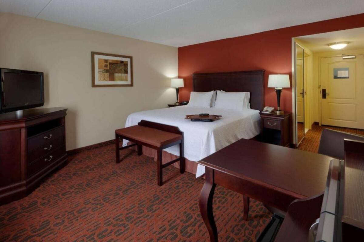 Habitación de hotel con cama, escritorio, silla, televisor, mesita de noche, lámpara y puerta.