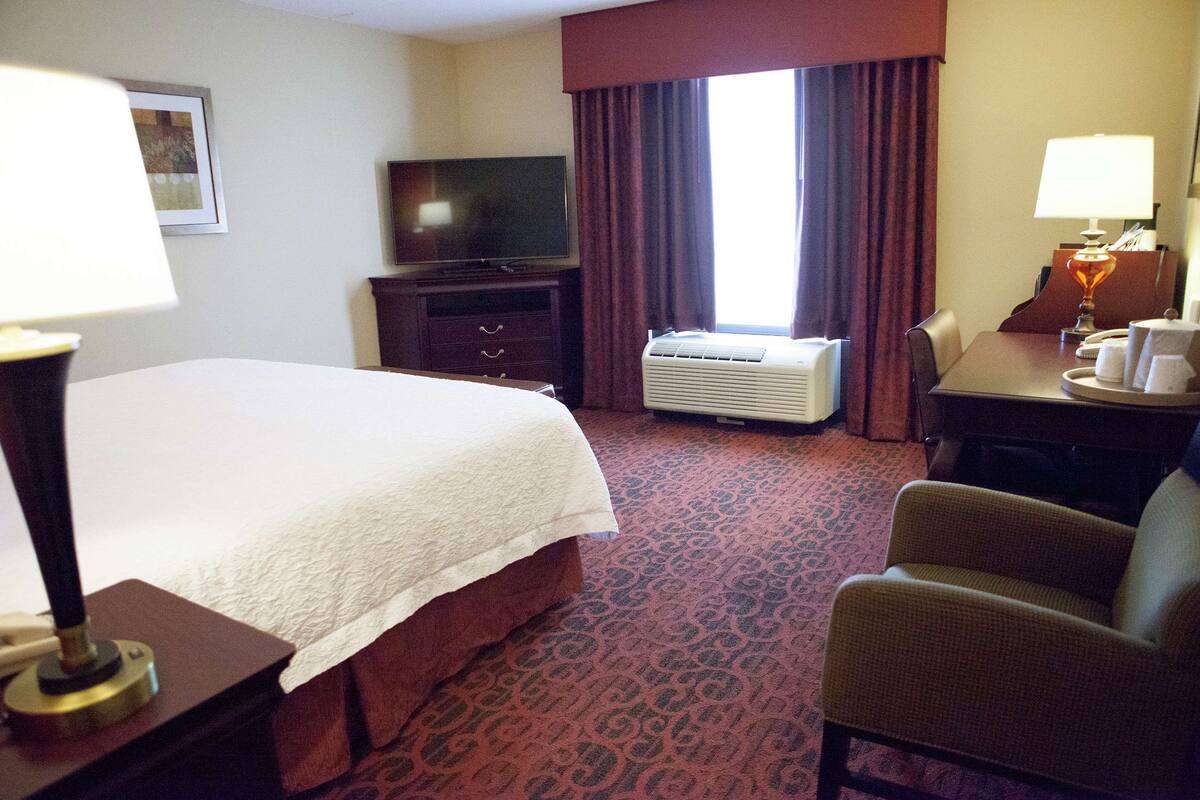 Habitación de hotel con cama, escritorio, silla, televisor, aire acondicionado y ventana con cortinas.