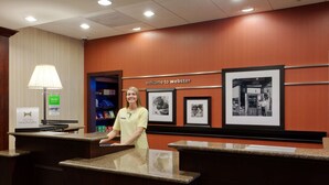 A hotel reception desk with a "Welcome to Webster" sign, framed black and white photographs on the wall, and a Hilton Honors sign on the counter. - Hampton Inn Rochester Webster (Webster)