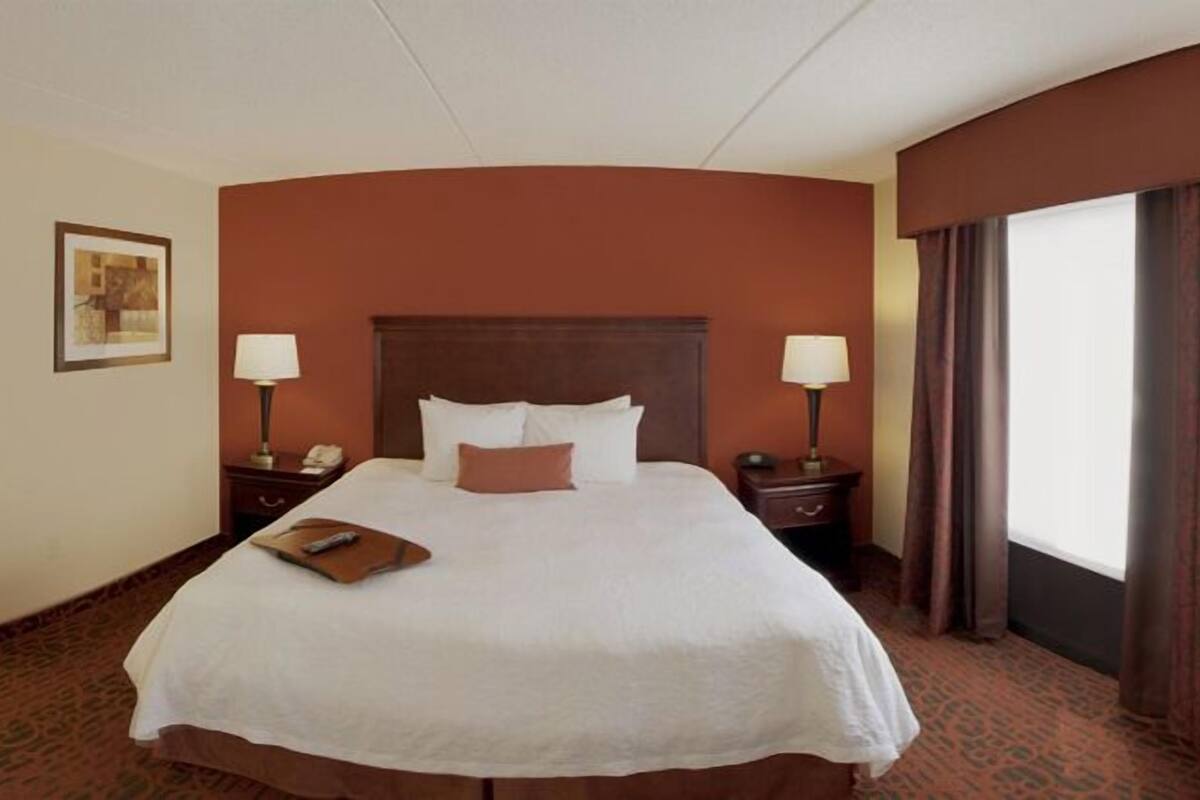 Habitación de hotel con una cama grande, dos mesitas de noche, una lámpara y un cuadro en la pared, además de un ventanal con cortinas.