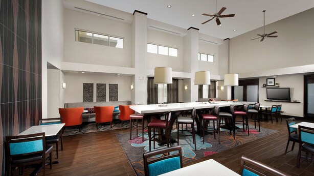 A spacious hotel lobby with a long communal table, multiple seating areas, ceiling fans, and a large flat-screen TV. - Hampton Inn & Suites Chicago Southland-Matteson (Matteson)
