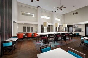 A spacious hotel lobby with a long communal table, multiple seating areas, ceiling fans, and a large flat-screen TV. - Hampton Inn & Suites Chicago Southland-Matteson (Matteson)