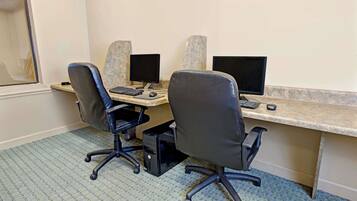 A room with two desks, each with a computer monitor, keyboard, and mouse. There are two office chairs and a small drawer under one desk. The room has a patterned carpet and a plain wall.