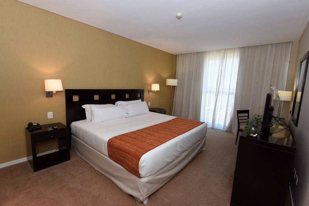 A hotel room with a large bed, two bedside tables, a desk with a chair, and a large window with curtains.