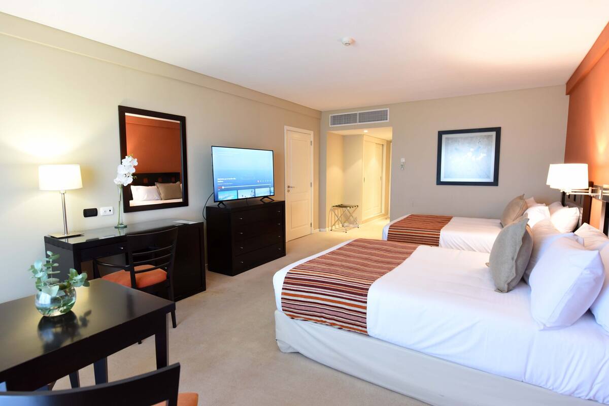 A hotel room with two beds, a desk, a TV, and a view of a bridge.