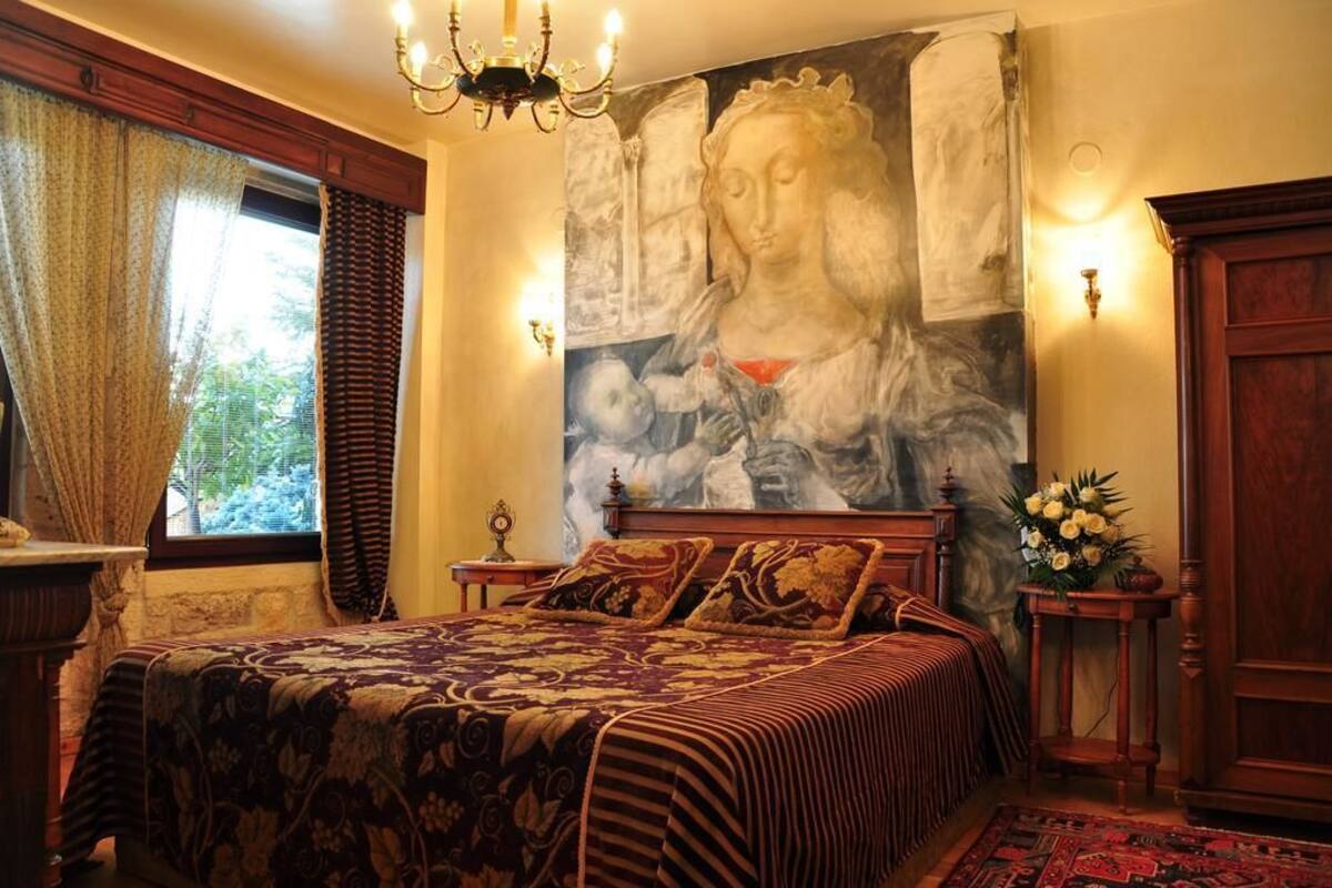 A bedroom with a large bed, a chandelier, a painting, and a window with curtains.