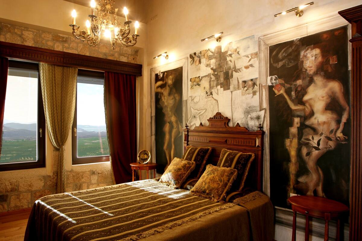 A bedroom with a large bed, a chandelier, a view through a window, and a painting on the wall.