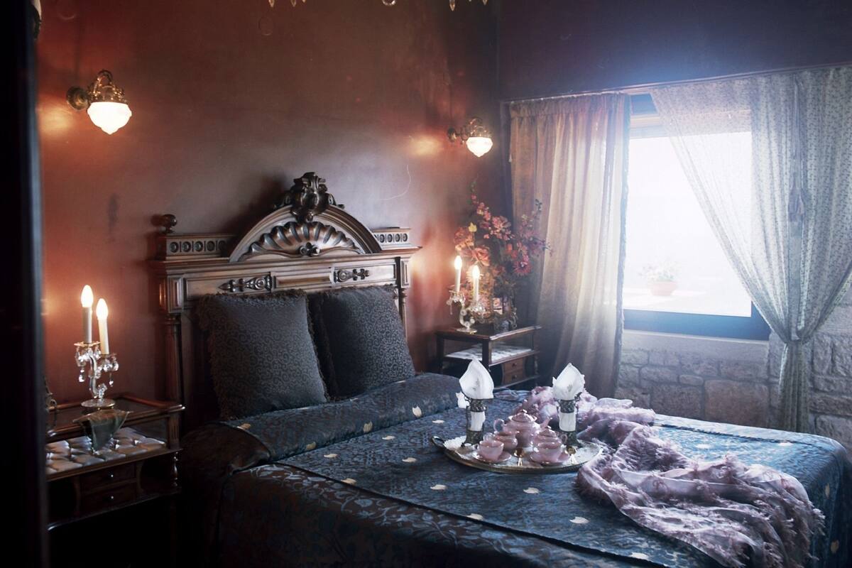 A bedroom with a large bed, a chandelier, a nightstand with a candelabra, and a window with curtains.