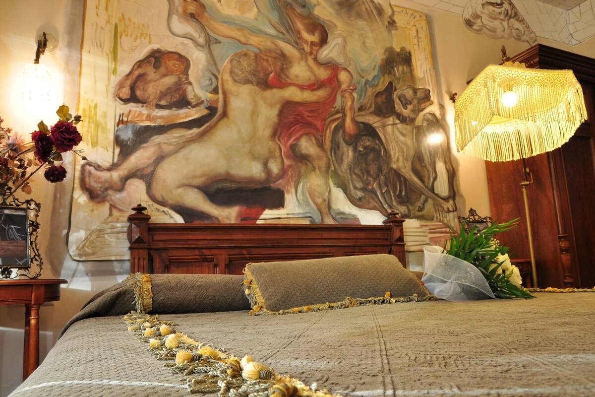A bedroom with a large painting, a bed with a beige bedspread, a wooden headboard, and a hanging light fixture.