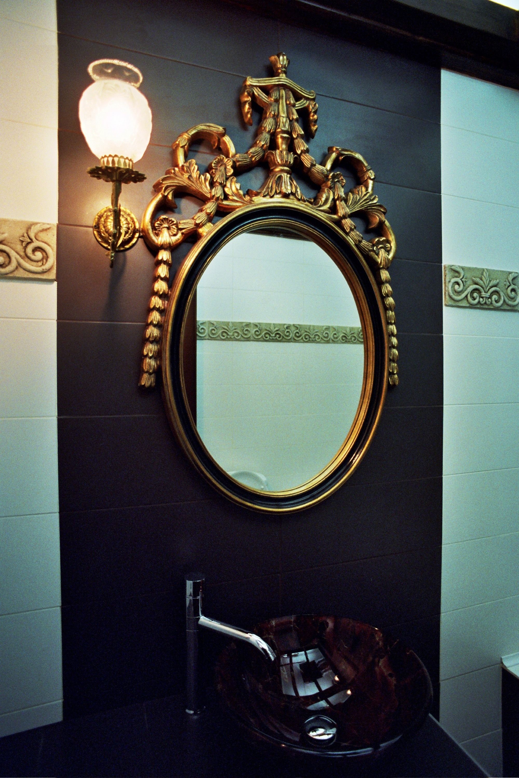 A bathroom with a round mirror, ornate gold frame, and a wall-mounted light fixture.