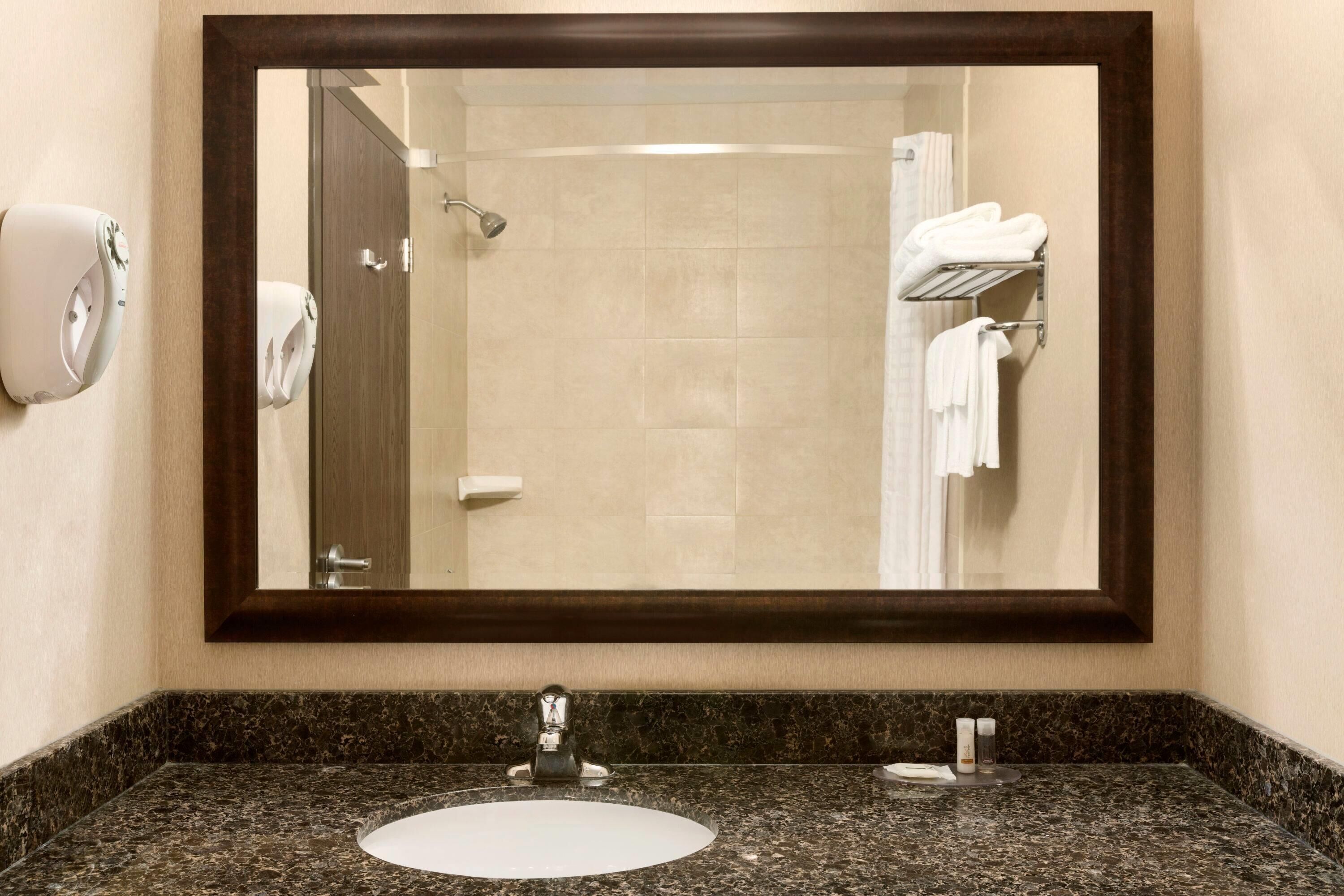 A bathroom with a large mirror, a sink, a towel dispenser, and a shower area.