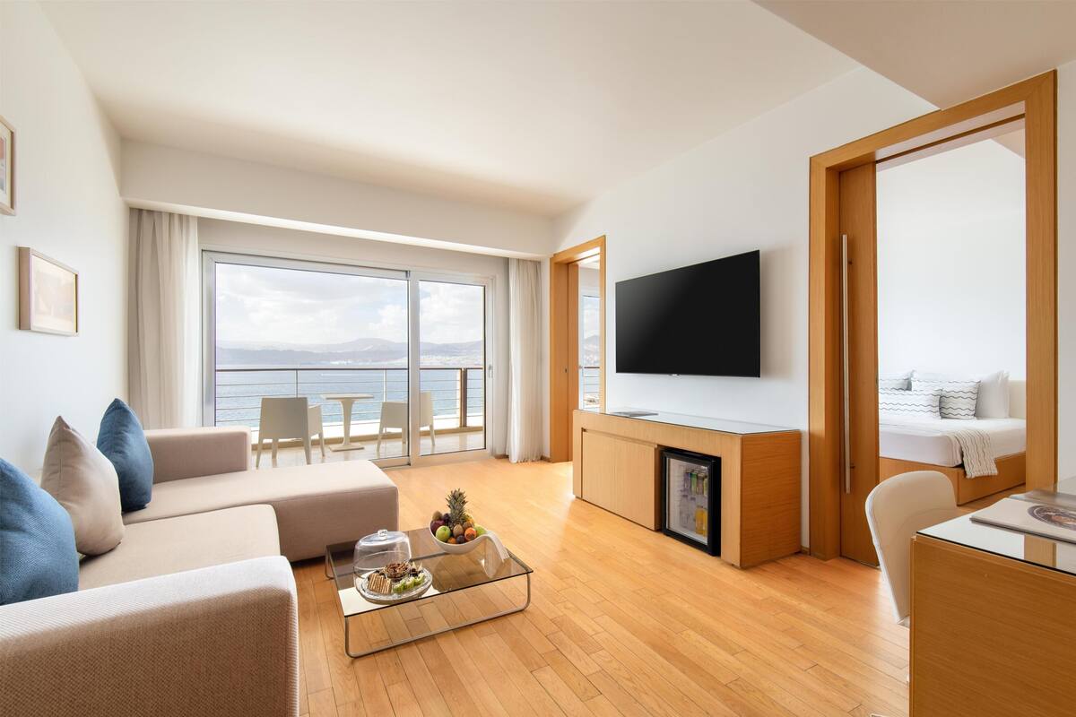 A modern living room with a sofa, coffee table, and a flat-screen TV. There's a balcony with a view of the sea.