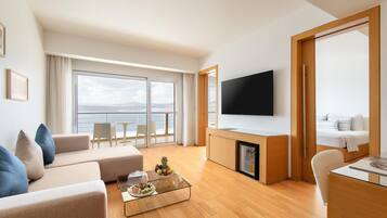 A modern living room with a sofa, coffee table, and a flat-screen TV. There's a balcony with a view of the sea.