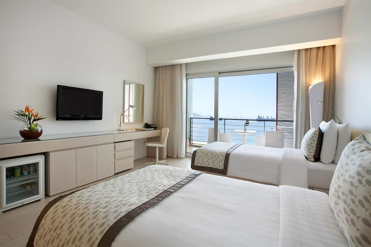 A hotel room with a large bed, a desk with a TV, a chair, and a view of the sea.