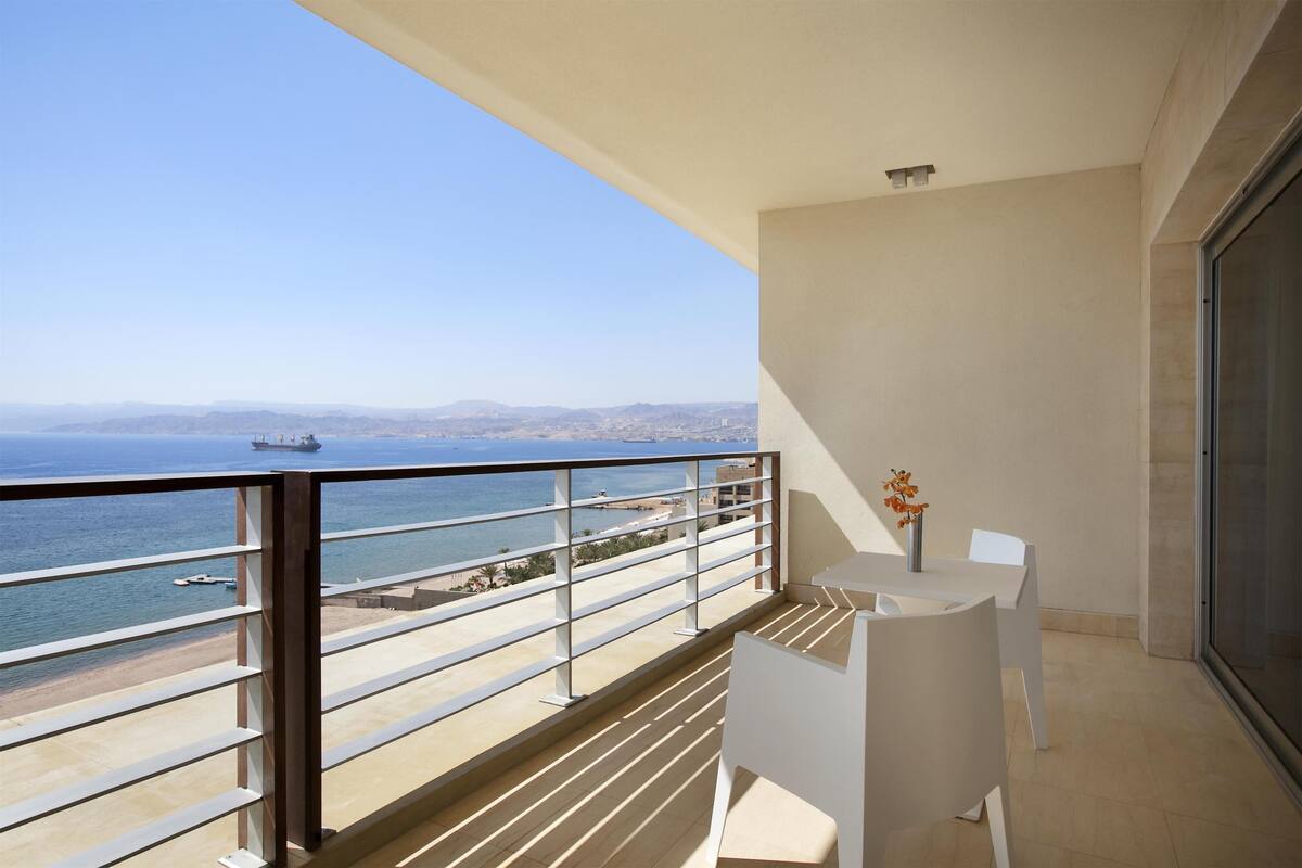A balcony with a view of the sea and a ship, white chairs, and a vase with flowers.