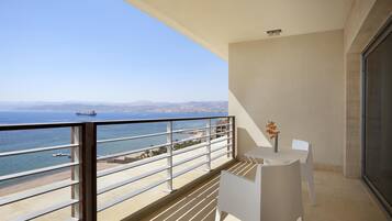 A balcony with a view of the sea and a ship, white chairs, and a vase with flowers.