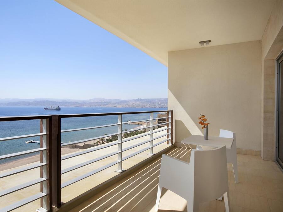A balcony with a view of the sea and a ship, white chairs, and a vase with flowers.
