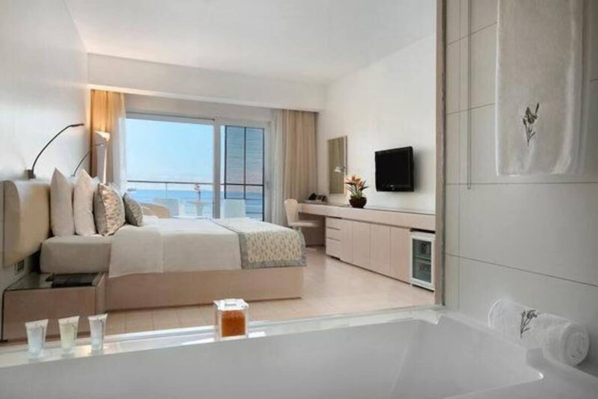 A modern hotel room with a large bed, a TV, a bathroom with a bathtub, and a view of the sea.