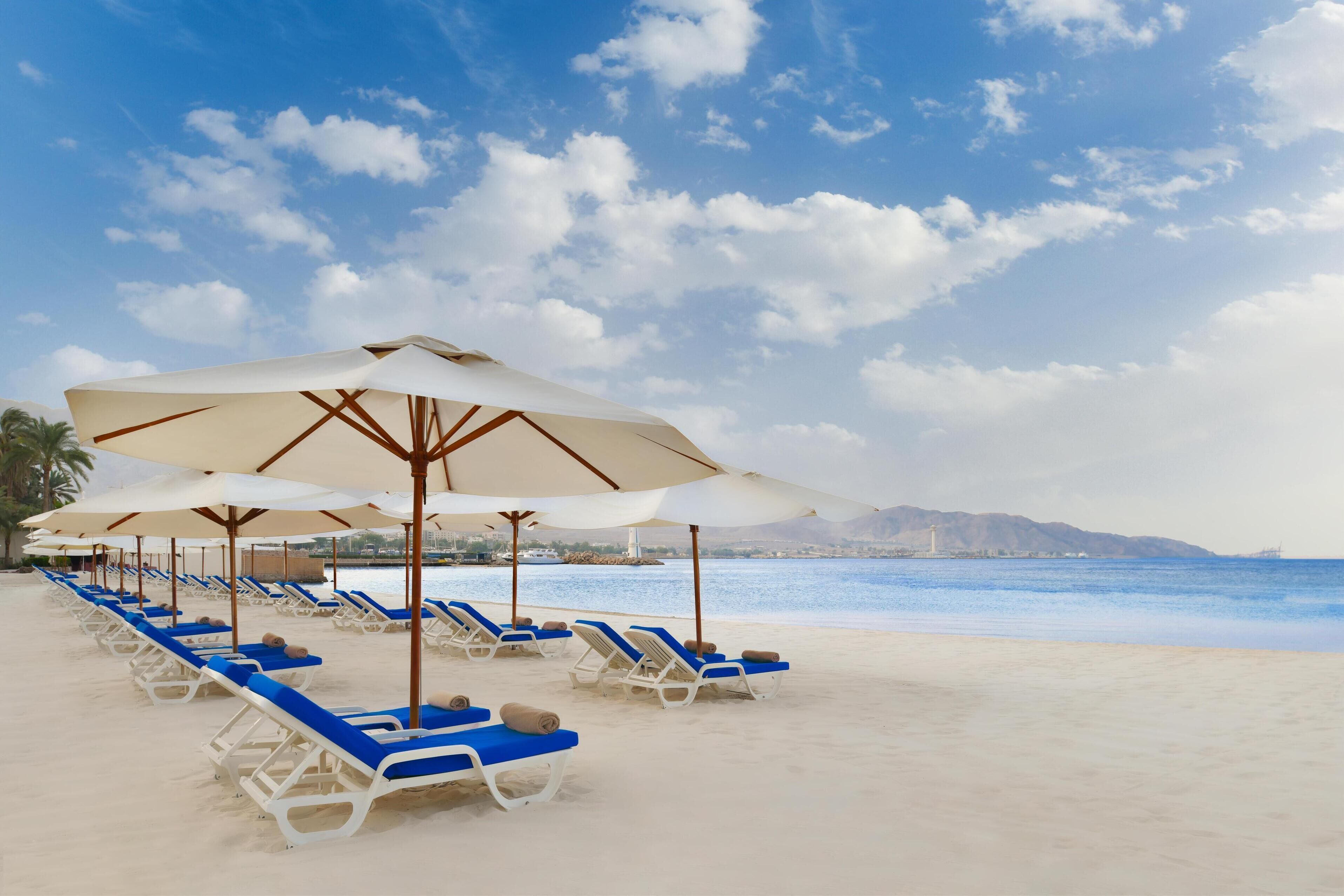 A beach with rows of sun loungers and umbrellas, a clear sky, and a calm sea.