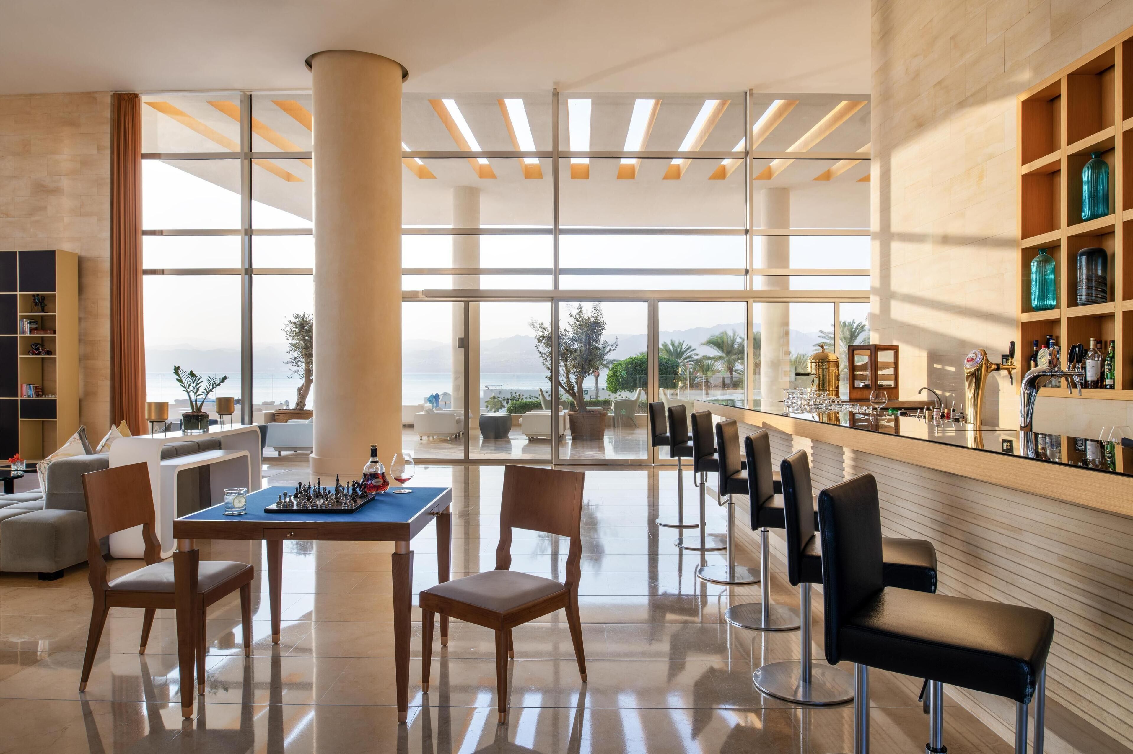 A modern hotel lobby with a large window offering a view of the mountains, a bar area with seating, and a chess table.