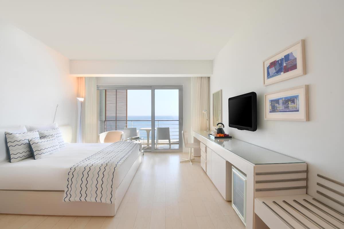 A modern hotel room with a bed, a desk, a TV, and a balcony with a view of the sea.