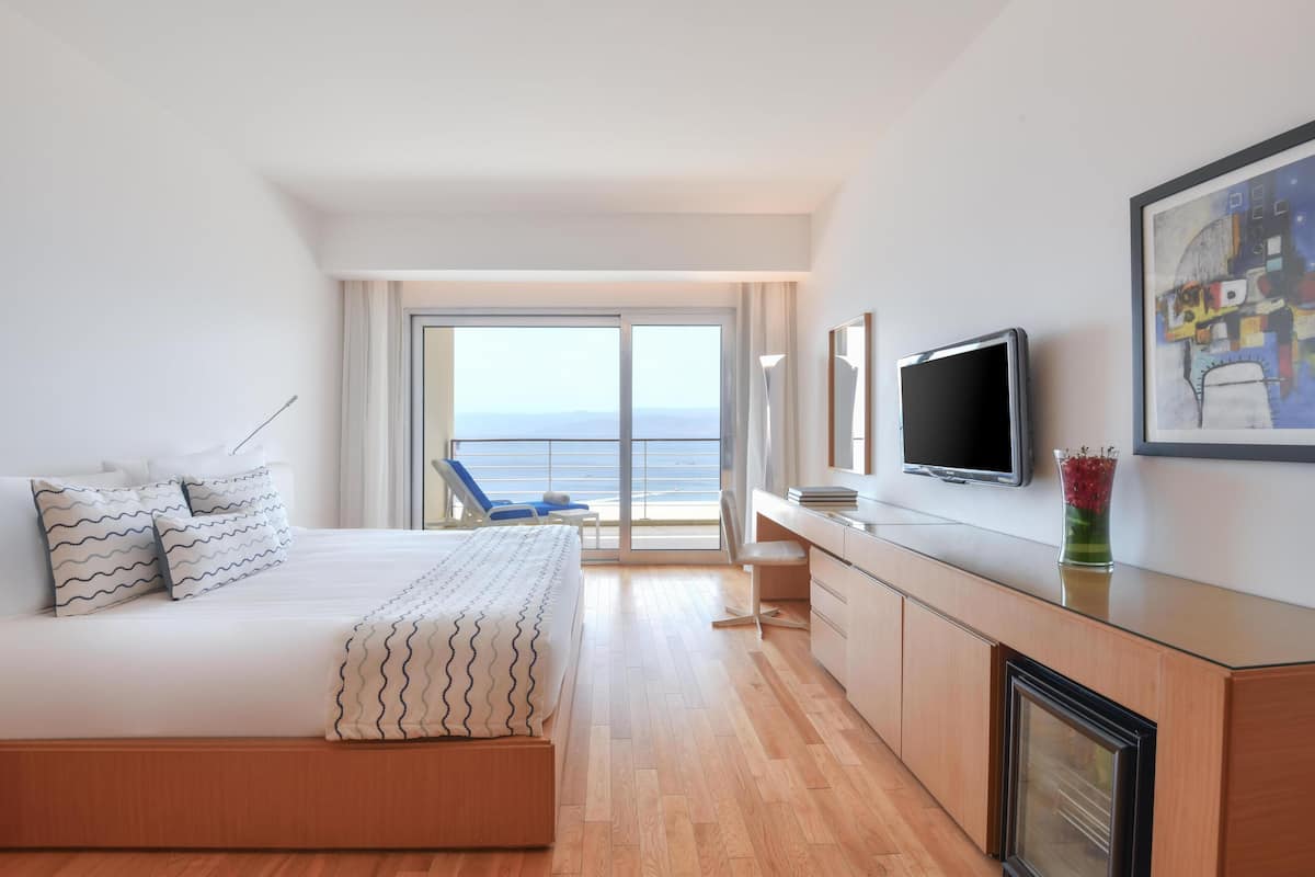 A modern hotel room with a large bed, a desk, a television, and a balcony with a view of the sea.