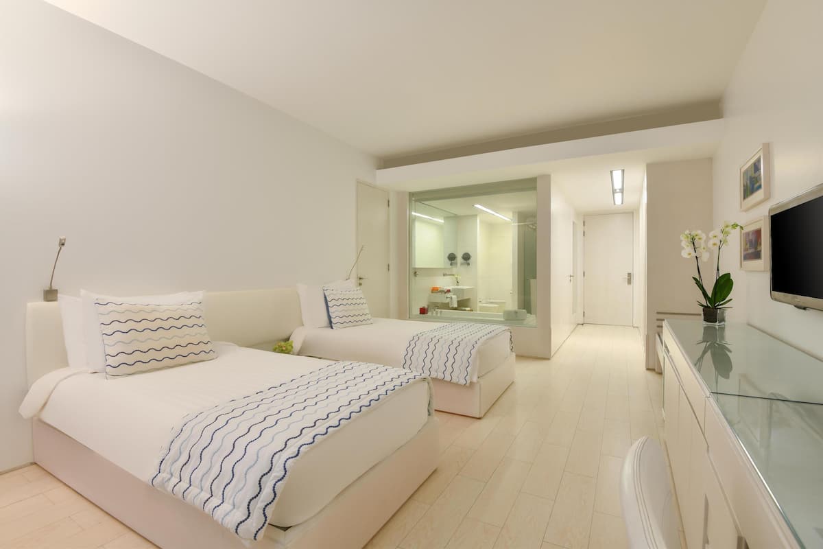 A modern hotel room with two beds, a glass cabinet, a TV, and a bathroom visible through a glass door.