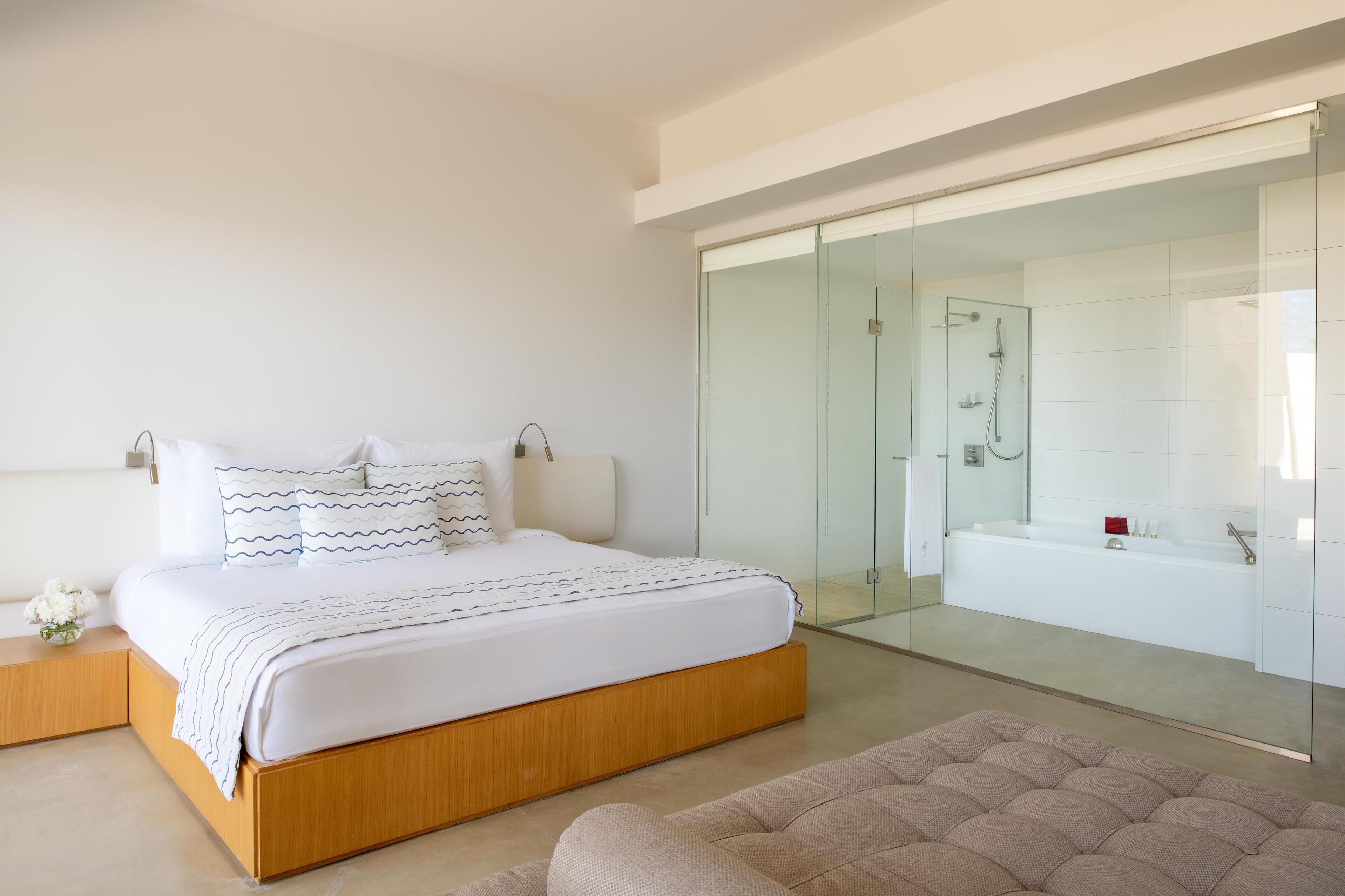 A modern bedroom with a large bed, a glass-enclosed bathroom, and a cozy seating area.