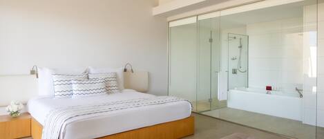 A modern bedroom with a large bed, a glass-enclosed bathroom, and a cozy seating area.