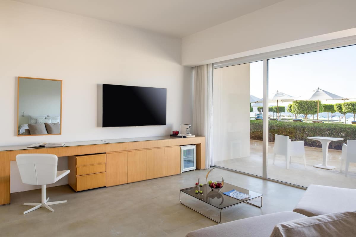 A modern hotel room with a large window, a flat-screen TV, a wooden desk, a white chair, and a glass coffee table.