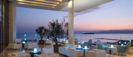 Un área de descanso moderna con sofás y sillones blancos, un gran árbol en maceta y vistas al mar al atardecer.