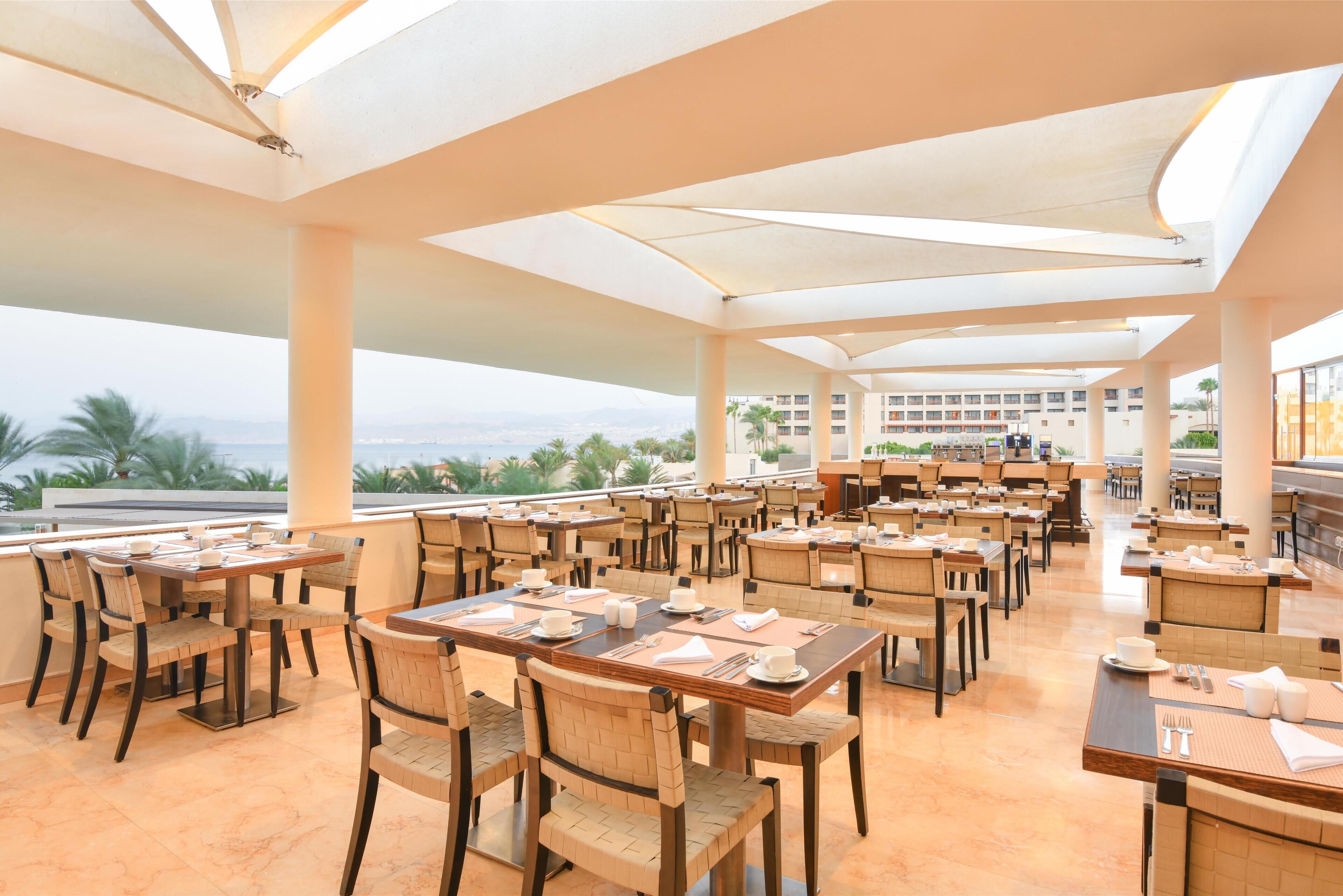 A spacious outdoor dining area with multiple tables and chairs, large umbrellas, and a view of the ocean and palm trees.