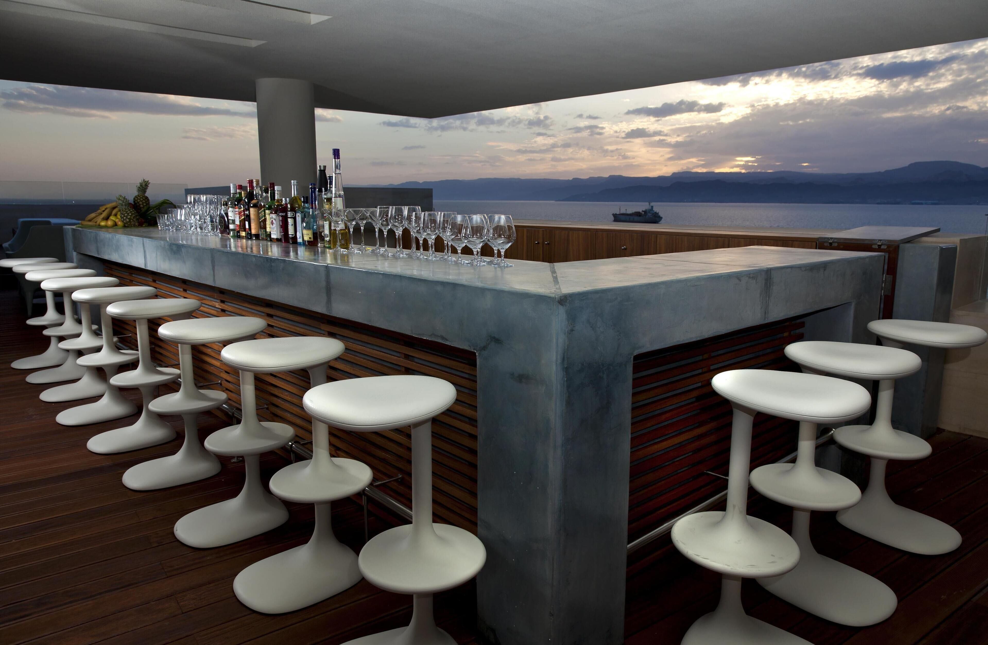 A bar with a view of the sea, wooden flooring, and white stools.