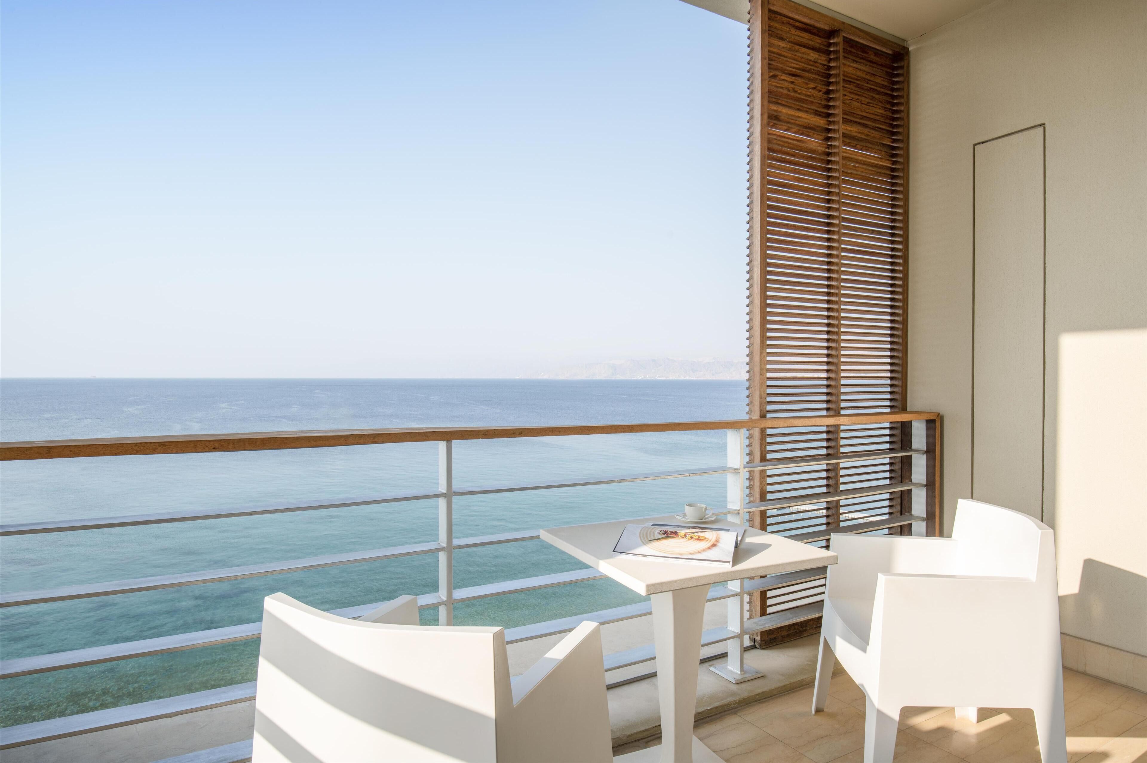 A balcony with a view of the sea, a table with a laptop and a cup, and white chairs.