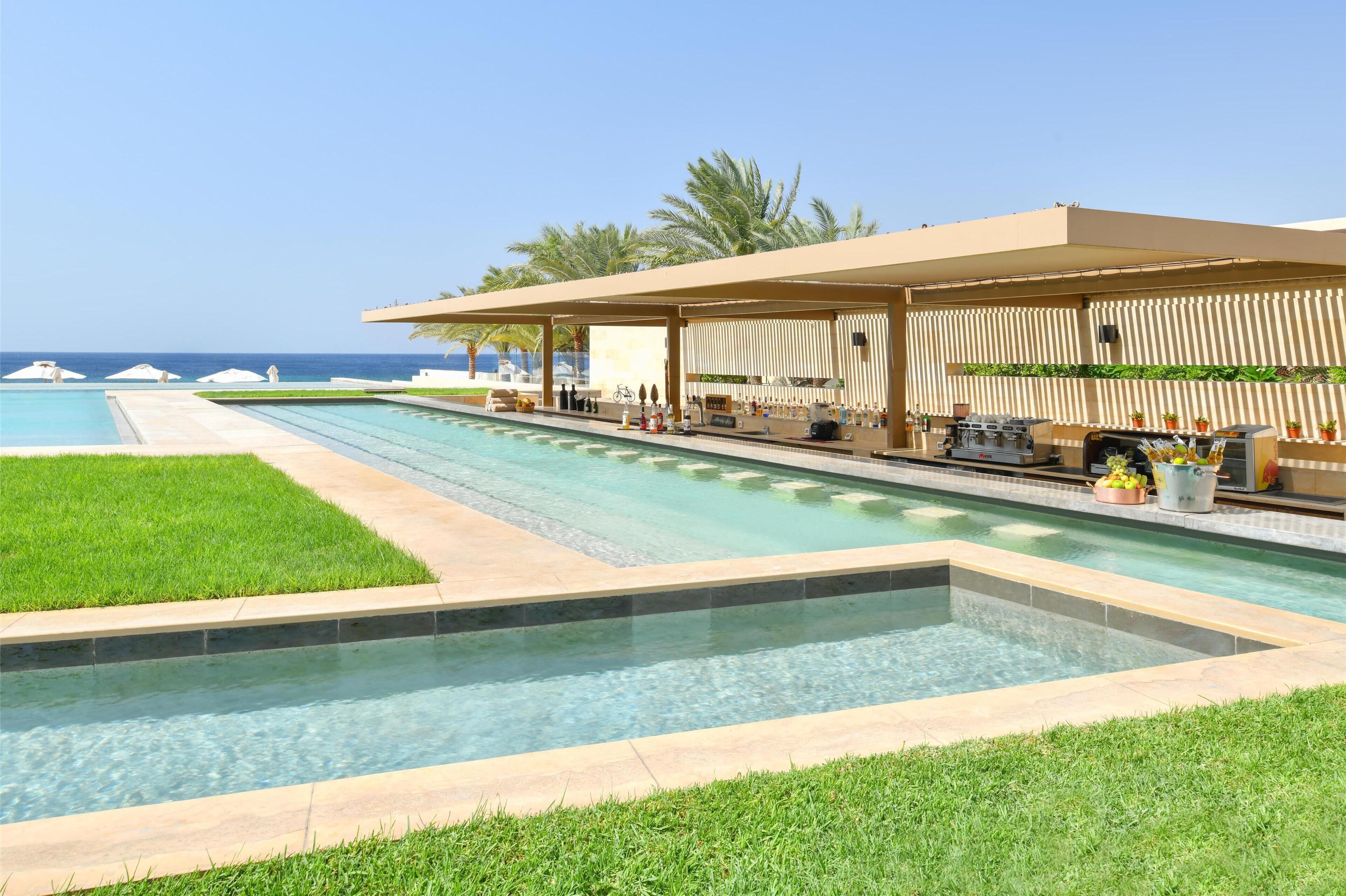Ein Poolbereich mit Bar und Sitzgelegenheiten, umgeben von Palmen und freiem Blick auf das Meer.