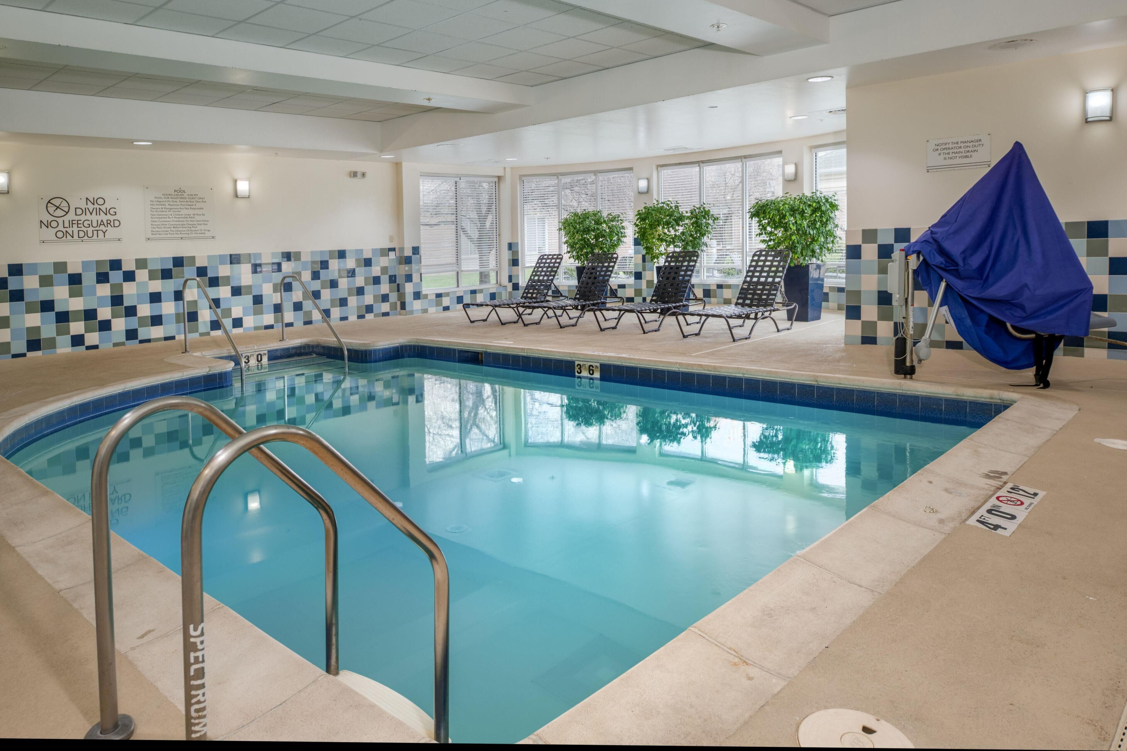 Piscina interna com água cristalina, placa de proibição de mergulho, cadeiras de espreguiçadeira e plantas em vasos.
