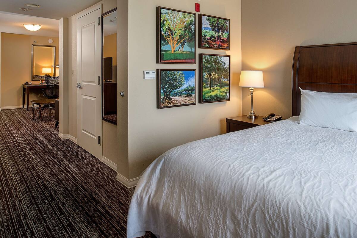 A hotel room with a bed, a desk with a chair, a lamp, and four framed pictures on the wall.
