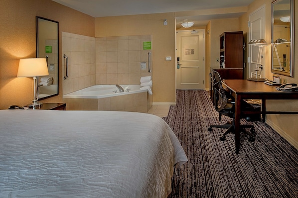 A hotel room with a large bed, a bathroom with a bathtub, a desk with a chair, and a desk lamp.