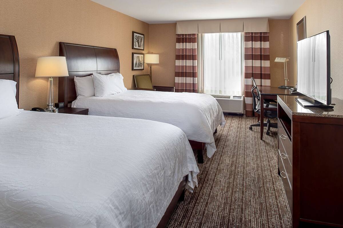 A hotel room with two beds, a desk, a chair, a television, and a window with curtains.