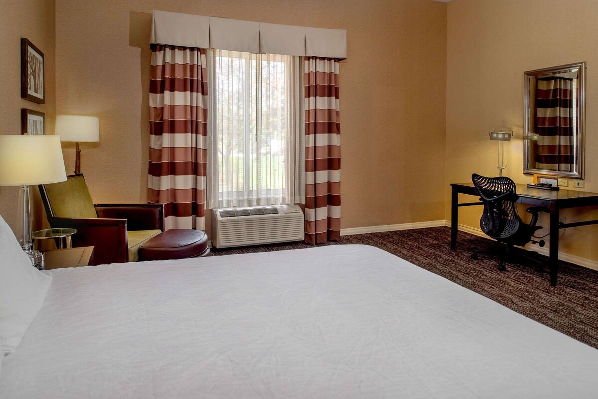 A hotel room with a large bed, a desk with a chair, a lamp, a window with striped curtains, and an air conditioning unit.