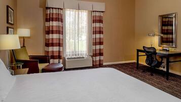 A hotel room with a large bed, a desk with a chair, a lamp, a window with striped curtains, and an air conditioning unit.