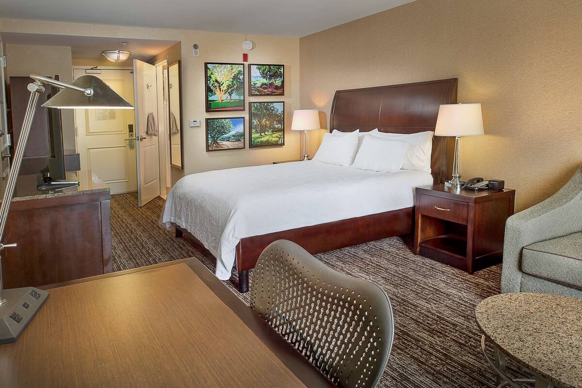A hotel room with a large bed, two bedside tables with lamps, a desk, a chair, and a carpeted floor.