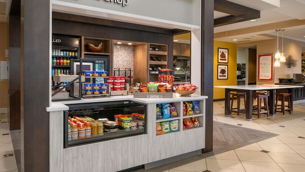 A snack kiosk with a variety of packaged foods and beverages, a cash register, and a sign reading 'The Shop.' - Hilton Garden Inn St. Louis Airport (St. Louis)