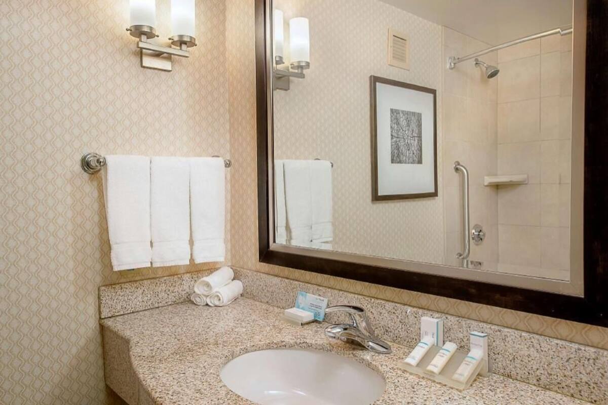A bathroom with a granite countertop, a sink, a mirror, two towels, a framed picture, and a shower.