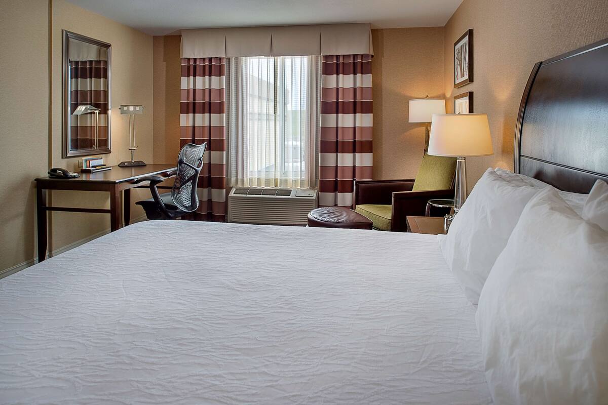A hotel room with a large bed, a desk with a telephone, a chair, a small table, a lamp, and a window with curtains.