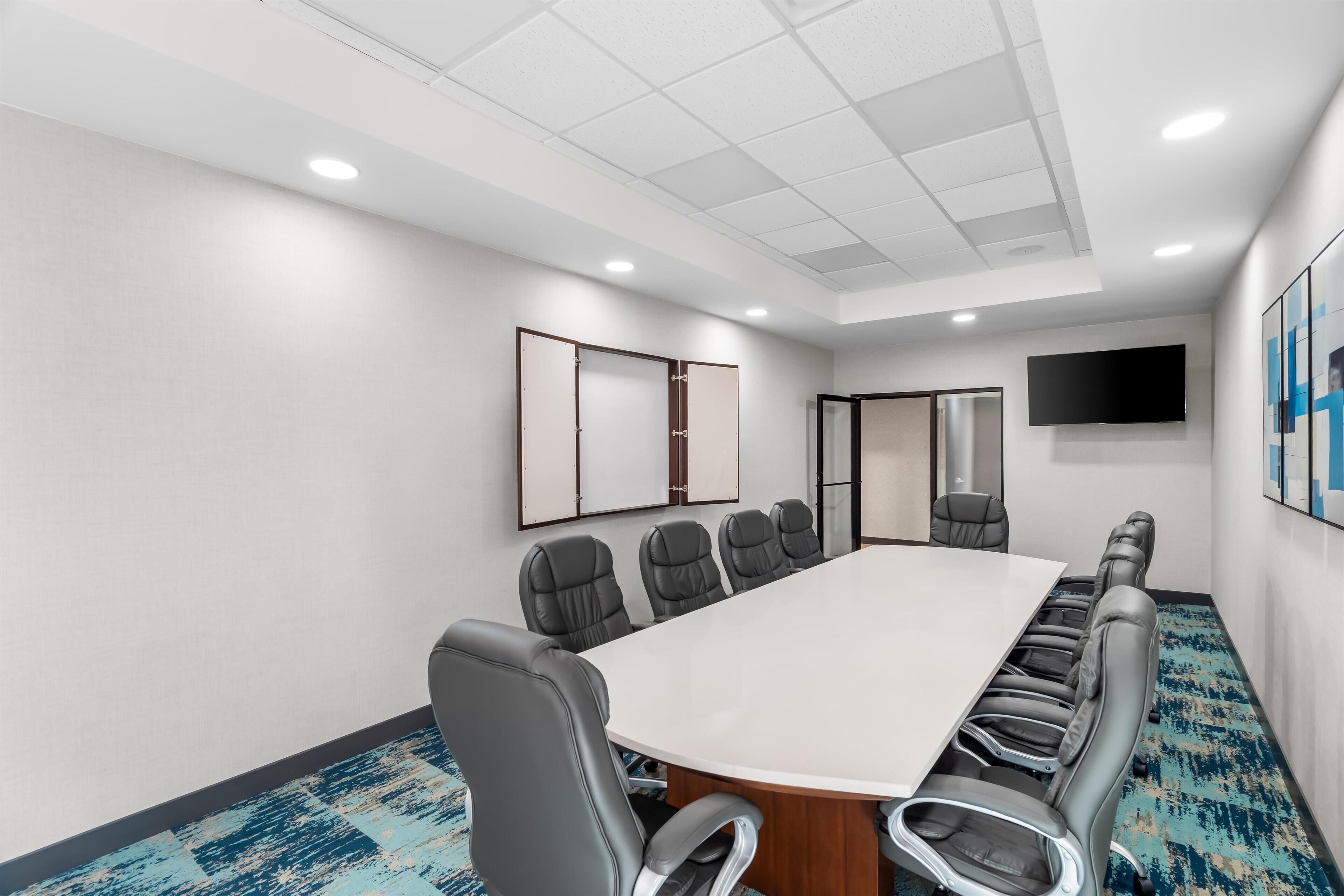 A conference room with a long table, multiple chairs, a flat-screen TV, and a large mirror on the wall.