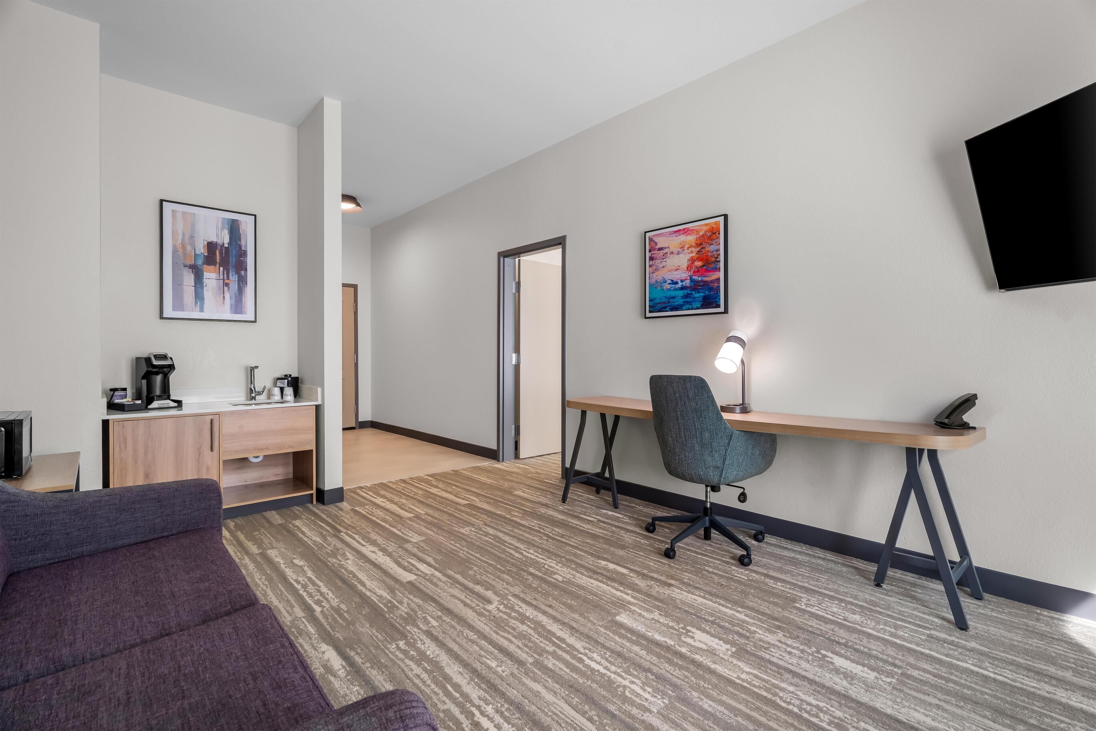 A modern hotel room with a sofa, desk, and chair, a flat-screen TV, and two framed artworks on the walls.