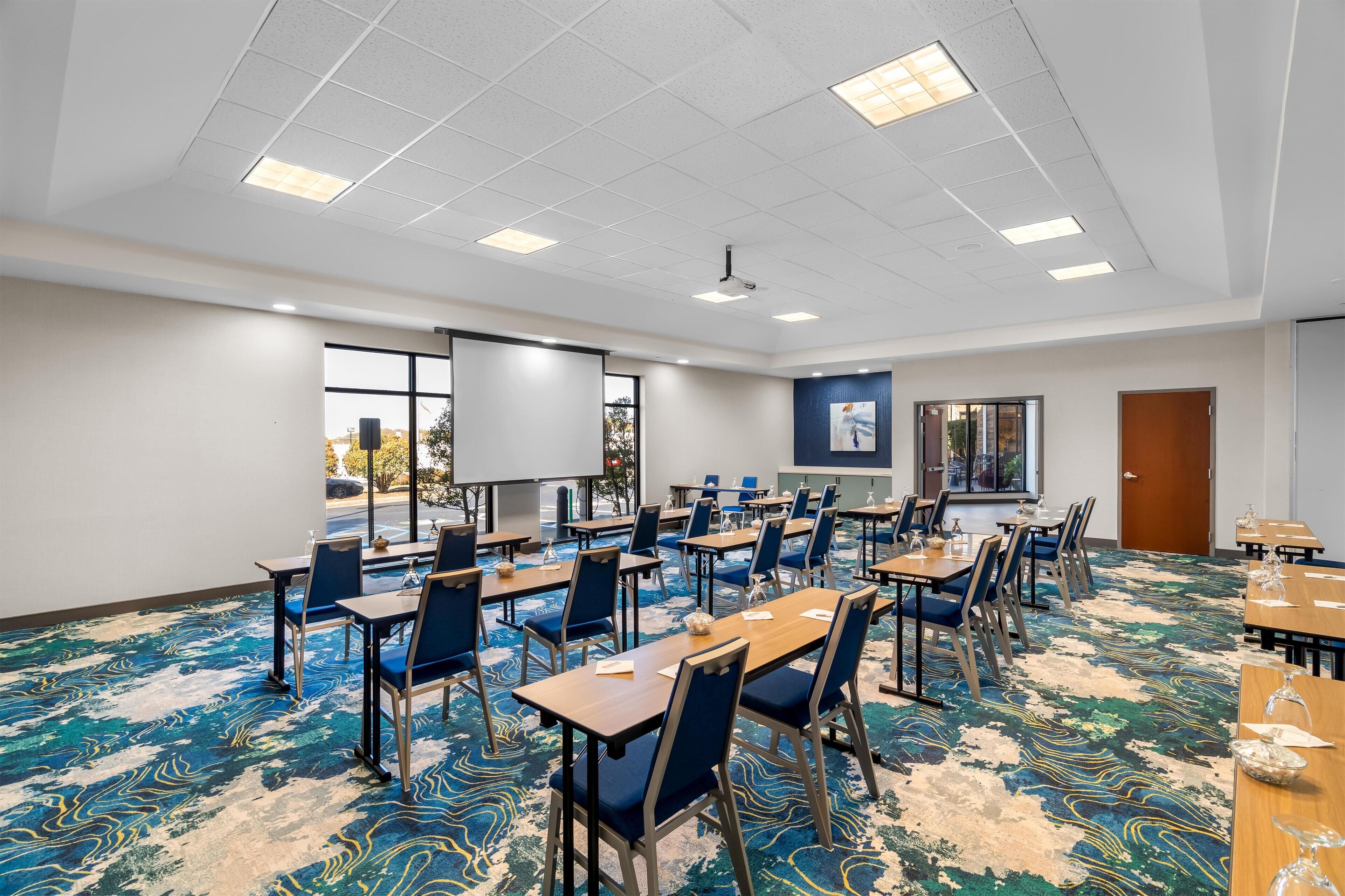 A conference room with tables, chairs, a projector screen, and a large window.