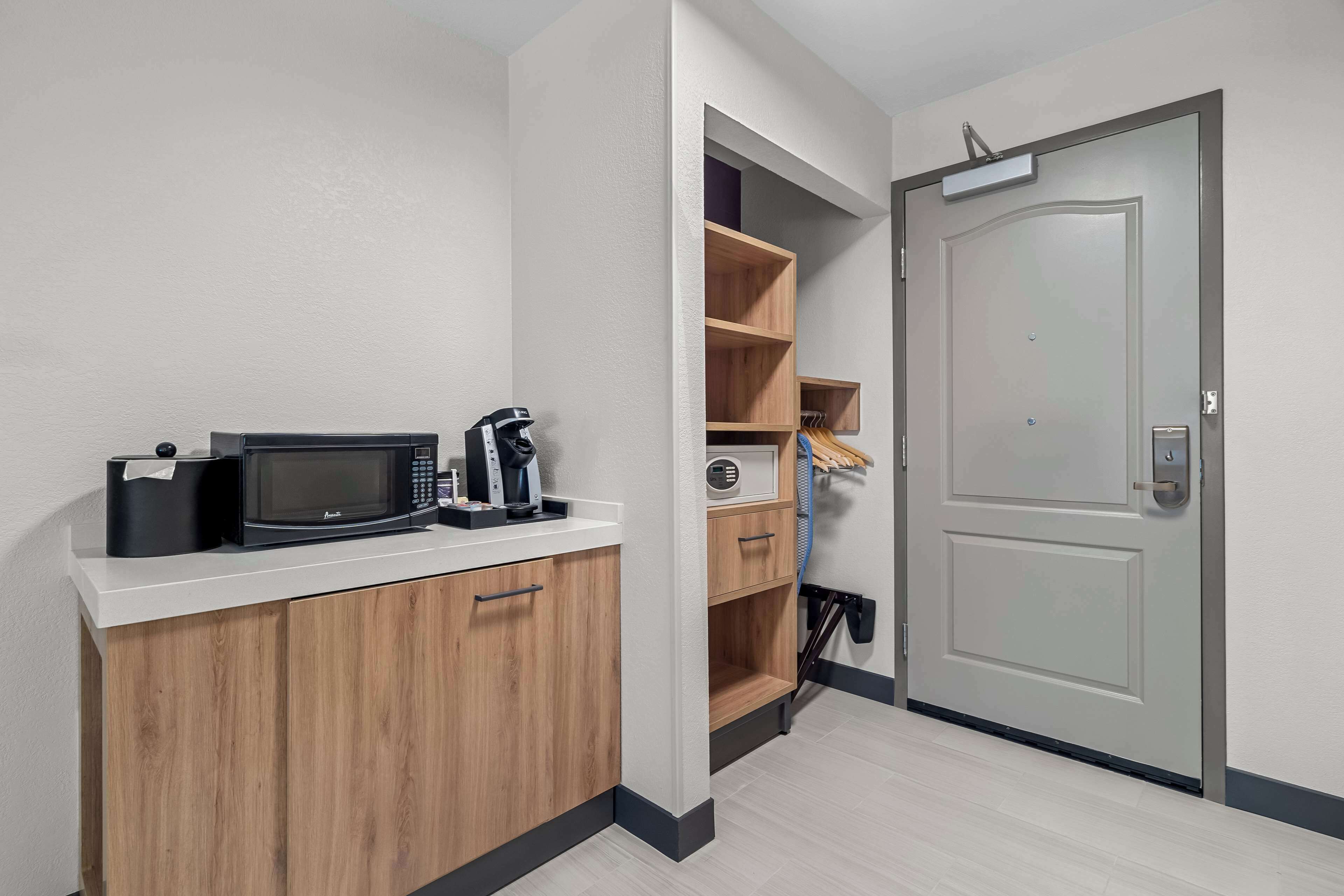 A kitchenette with a microwave, coffee maker, and storage cabinets. There is a door with a handle and a small shelf above it.